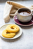Close-up of a warm coffee cup on a decorative plate, paired with golden shortbread biscuits on a small bowl, set on a textured table with a linen napkin for a cosy breakfast atmosphere