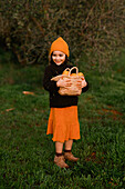 A smiling little girl wearing a cap and an orange skirt holds a basket of oranges in a lush green field. Her autumn clothes reflect the bright colours of the season.