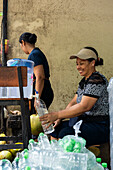 Two Asian women prepare fresh coconut water from bottles and a jug in a rustic setting. Surrounded by coconuts and plastic bottles, they work diligently and smile at each other.