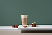 A tall glass of coffee with milk cream and brown sugar stands elegantly on a minimalist surface, surrounded by coffee capsules and a spoon, against a muted background