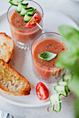 Two glasses of gazpacho with cucumber and tomato skewers, served with toasted bread on a white plate Perfect for a light, refreshing starter or snack