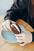 Close-up of hands holding a chocolate cake with a creamy filling on a light-coloured ceramic plate on a wooden table to capture a sweet moment.