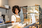 A café employee with vitiligo in a casual brown apron smiles from behind a glass patisserie display.