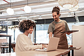 A barista in an apron serves a smiling customer with a laptop in a cosy café The warm atmosphere is enhanced by the stylish lighting and modern decor in the background