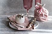 Beautifully arranged Easter table with a vase made from a broken eggshell with feathers, surrounded by pink elements and spring decorations. Soft light emphasises the cheerful mood.