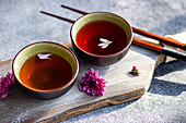 Two cups of tea with delicate petals float on the surface. They stand on a wooden board, surrounded by purple flowers and sticks, creating a calm and inviting ambience.