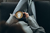 Eine Person in einem gemütlichen Pullover genießt einen wunderschön zubereiteten Milchkaffee mit komplizierter Schaumkunst, während sie bequem mit gekreuzten Beinen auf einem weichen Stuhl sitzt und einen heiteren Kaffeemoment festhält