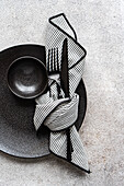 A minimalist black and white placemat with a plain black plate, bowl and cutlery, complemented by a striped napkin. Perfect for elegant table settings.