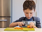 A focussed young child in a blue apron cuts a carrot on a green chopping board. The picture shows the child's concentration and dexterity in the kitchen.