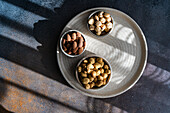 Top view of a round plate with three bowls filled with almonds, hazelnuts and peanuts, which are beautifully emphasised by natural sunlight and shadows.