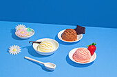 Colourful ice cream scoops in white bowls against a strong blue background, decorated with flowers, chocolate and a strawberry.