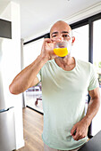 A man in a casual T-shirt sips a refreshing glass of orange juice at home. The light and airy kitchen environment emphasises the peaceful morning moment and promotes relaxation.