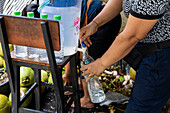 A woman fills plastic bottles with fresh coconut water from a vending machine at a street market. The picture shows bottled coconut water, coconuts and an insight into tropical street culture.