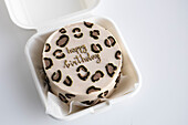 Top view of a stylish bento birthday cake with a leopard print and a golden happy birthday message, served in a white box to take away.