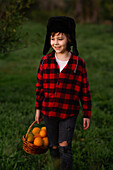 A smiling child in a red chequered shirt and a winter cap holds a basket of oranges in his hand as he walks across a green meadow. The picture captures a moment of joy and harvest outdoors.