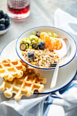 A nutritious breakfast bowl with muesli, yoghurt, blueberries, kiwi and mandarin orange slices, accompanied by crispy waffles, served on a white plate with a spoon.