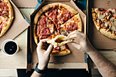 Hands cut from above of an unrecognisable person holding a slice of pizza, with several open pizza boxes with different toppings visible. The shot suggests a casual, friendly meal.