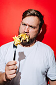 A bearded man in a white T-shirt playfully holds French fries in his mouth while making a humorous facial expression. The bold red background provides a striking contrast to the quirky and comedic food moment.