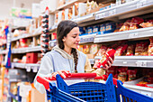 A woman with a pigtail looks at a product in a supermarket aisle while pushing a shopping trolley.