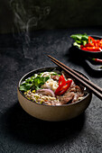 A steaming bowl of Vietnamese beef pho noodle soup (Pho Bo), garnished with slices of beef, fresh herbs, onions and a slice of red chilli, artfully presented against a dark, textured background.