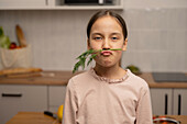 A young girl playfully holds a dill stalk under her nose, imitating a moustache. She is standing in a kitchen with a bag of vegetables in the background and has a humorous expression on her face.