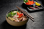 Traditional Vietnamese beef pho noodle soup in a ceramic bowl, garnished with slices of beef, herbs, red chilli and lime on a dark stone background.