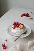 Beautifully arranged redcurrant and passion fruit pavlova on a white ceramic plate on a table covered with fabrics. The dessert is garnished with fresh redcurrants and a sliced passion fruit, creating a visually appealing and appetising picture.