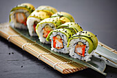 Delicious sushi rolls with avocado and black sesame seeds, presented on a bamboo mat against a dark slate background.