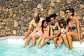 A cheerful family of five relaxes by the pool on a sunny day, laughing and enjoying their holiday. The background is a rustic stone wall that adds to the atmosphere.