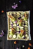 A tart with courgettes and edible flowers is beautifully arranged on a dark wooden table. The bright colours of the flowers contrast with the green courgettes and create a visually appealing dish.