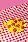 A colourful flower arrangement with yellow and orange flowers on a yellow plate, placed on a red and white checked tablecloth against a pink background, creates a lively contrast.