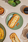 A delicious slice of toast, topped with peanut butter and banana slices on a green plate. Served with a cup of coffee, almonds and a small plant for a cosy breakfast situation.