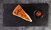 A delicious slice of cheesecake with a golden brown surface, served on a slate plate, accompanied by a rich berry sauce in a small black bowl. Perfect for dessert photos.