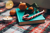 Cheerful Christmas scene with a burning candle, a candy cane and fresh mandarins on a blue-green book, all embedded in a colourful, textured blanket that conveys a feeling of Christmas warmth and joy.