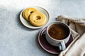 Close-up of a warm coffee cup on a decorative plate paired with golden shortbread biscuits on a small bowl, set on a textured table with a linen napkin for a cosy breakfast atmosphere
