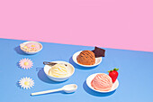 Various flavours of ice cream in white bowls, presented against a striking pink and blue background, accompanied by decorative flowers, a strawberry and a piece of chocolate.