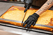 An artisan with gloves skilfully cuts a traditional Italian cake with a large knife The large baking tray and tattooed arm create an authentic bakery atmosphere