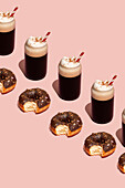Flat chocolate donut with removed bites, placed between coffee cups with whipped cream and red and white straws against a pastel pink background