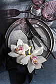 A sophisticated table setting with a textured plate, dark-coloured cutlery and elegant white orchids. Two purple glasses add a touch of colour to this sophisticated arrangement.