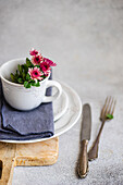 Elegantly laid table with a white cup with pink flowers on a napkin, on stacked plates and accompanied by old silver cutlery on a wooden board
