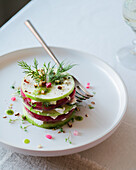 Top view of a finely composed gourmet salad with cucumbers, radishes and feta cheese, garnished with dill, chopped nuts and edible flowers on a white plate.