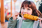 A young girl enjoys a spiral-shaped tornado potato on a spit at an outdoor market. The golden, crispy snack emphasises the festive atmosphere and creates joy and excitement.