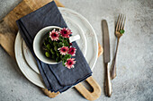 On a stylishly laid table, a white cup with pink flowers stands on a plate with a grey napkin, complemented by vintage cutlery on a rustic wooden board