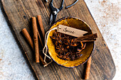 Top view of a rustic, textured wooden surface with star anise and cinnamon sticks in a glazed ceramic bowl labelled Spices. Scissors and a scattering of spices add charm and detail to the composition.