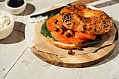 Ein Gourmet-Bagel mit geräuchertem Lachs, Kapern und frischem Grün auf einem Holztablett. Die Sesamkörner verleihen dem Bagel Struktur und machen ihn zu einer köstlichen, schmackhaften Frühstücksvariante.