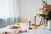 An elegantly laid table with refreshing tangerine drinks, burning beeswax candles, artistic floral arrangements and fine dining utensils. A cheerful, sophisticated ambience, perfect for a special occasion.