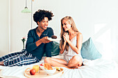 A young couple have a cosy breakfast in bed with juice, fruit and pastries. They smile and enjoy a relaxed morning together, creating a warm, cheerful atmosphere.