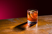 A classic whiskey old fashioned on a wooden table. The cocktail is served with ice cubes and a slice of dried orange, which casts a dramatic shadow and creates an inviting and sophisticated atmosphere.
