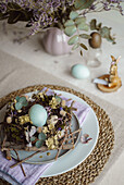 Top view of a table set for Easter with a plate featuring a rustic nest of twigs and naturally coloured eggs, surrounded by floral decorations and subtle tableware accents in soft, natural lighting.