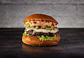 A gourmet burger with fresh salad, juicy beef, melted cheese, guacamole and a dollop of spicy sauce on a toasted brioche bun on a dark, rustic surface.